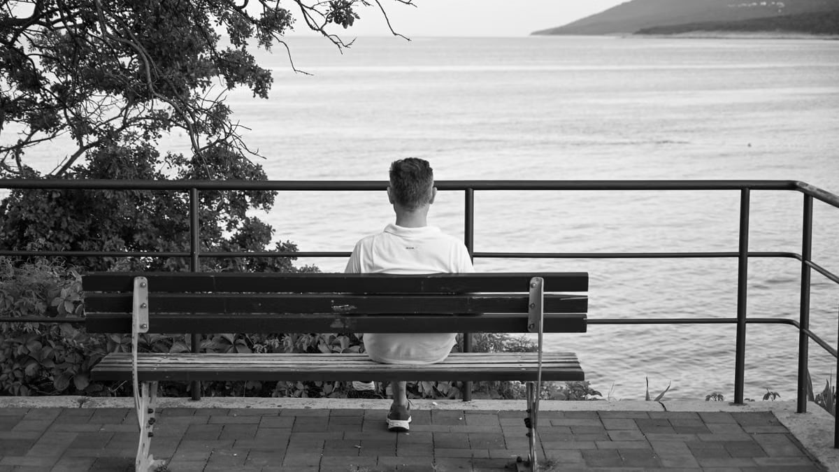 A man sits on a bench overlooking a tranquil sea, capturing a peaceful moment at the waterfront.